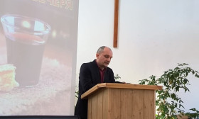 Man speaking at a church podium with a projection screen showing 'Lord's Supper' and a cross on the wall.