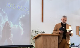 Man preaching from a wooden podium in a church, with a cross on the wall and a screen displaying 'PRAYER' in Russian.