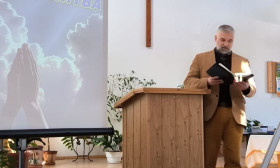 A man in a suit stands at a wooden podium, reading from a book with a cross on the wall behind him.