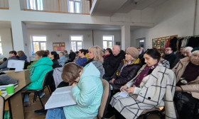 Diverse group of people attending a community meeting or lecture, focused and engaged in a bright, open room.