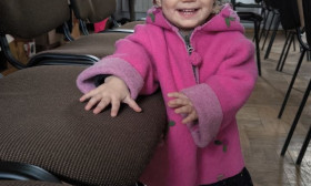 Adorable toddler girl in a pink coat and white hat, smiling brightly while standing next to chairs indoors.