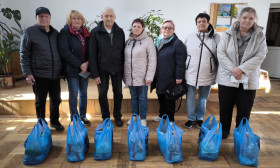 Seven elderly people stand behind a row of blue bags, likely containing humanitarian aid, in a community setting.