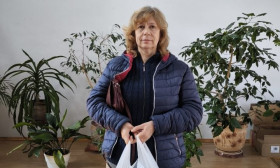 Woman in blue jacket holding a white bag, standing in a church with a wooden cross and potted plants.