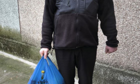 A man in a black jacket and pants holds a blue bag, standing on concrete against a textured wall.