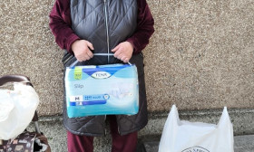 Elderly woman holding adult diapers, standing outdoors with a Church of Christ bag, symbolizing humanitarian aid.