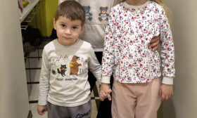 A mother and her two young children, a boy and a girl, stand in a hallway with two bags of groceries.