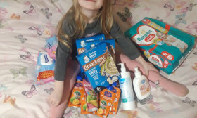 Happy young girl sitting on a bed surrounded by baby products, including diapers, baby food, and lotions.