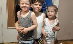 A smiling woman stands behind three young boys, who are proudly displaying two bags filled with groceries and essential supplies. The scene captures a moment of community support and aid.