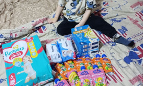 A young child sits on a bed with a Union Jack duvet, surrounded by baby products including diapers, wipes, and food pouches.