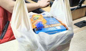 A white plastic bag filled with groceries, including baby products and food, sits on a wooden counter in a store.
