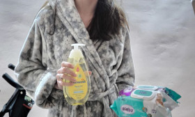 A young woman in a cozy bathrobe holds baby shampoo and a pack of baby wipes, ready for baby care.