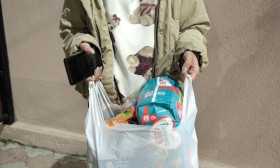 A woman stands holding a white plastic bag filled with groceries and baby diapers, wearing a teddy bear hoodie and slippers.