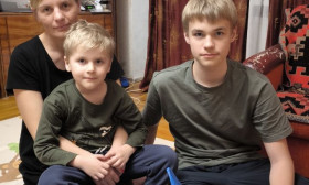 A family, a mother and her two sons, sit on the floor with a blue bag of groceries, looking at the camera.