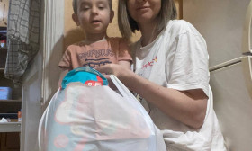 Mother and child with a large white bag of groceries, likely diapers and baby essentials, in a home setting.