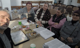 A group of five elderly women and one man are gathered around a table, studying books and enjoying refreshments.